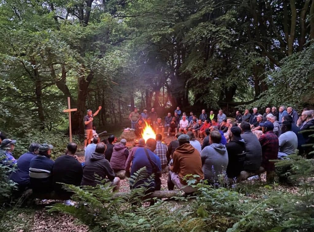 A men's evening community gathering around a campfire in the woods
